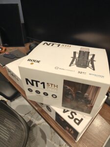 A boxed RØDE NT1 5th Generation studio condenser microphone sits on a wooden desk next to a computer monitor and headphones. The packaging highlights features like 32-bit float recording and dual USB/XLR connectivity, hinting at the recent launch of a professional podcast setup.