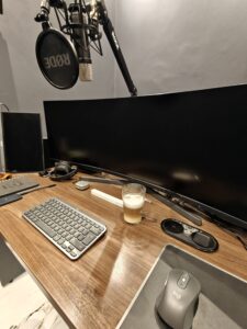 A podcast recording setup featuring a RØDE microphone on a boom arm, an ultra-wide monitor, a wireless keyboard and mouse, headphones, and a clear glass mug of coffee on a wooden desk.